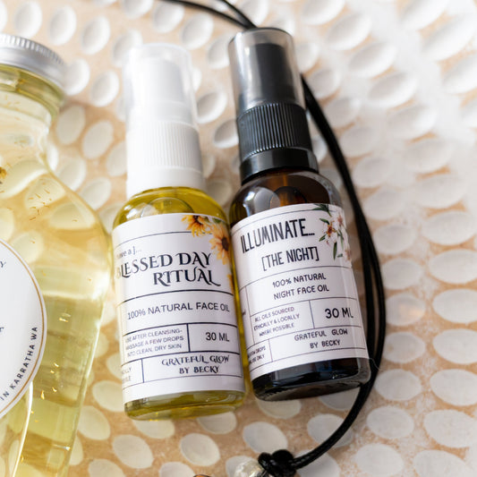A styled flat-lay of several small wellness and ritual products on a white textured plate, including a small bottle labeled Blessed Day Ritual, and a dark bottle labeled Illuminate (The Night).