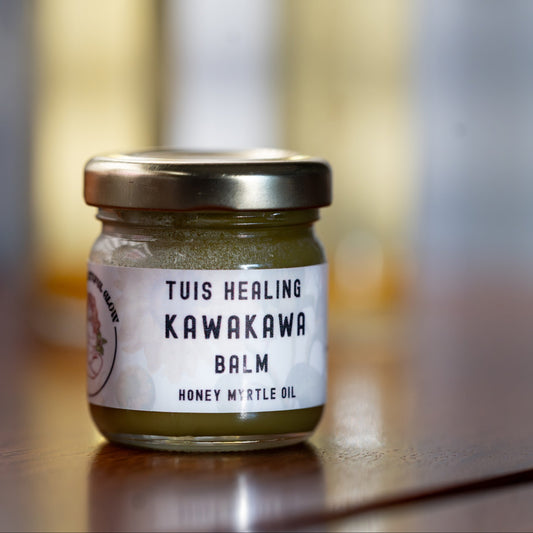 A small glass jar of Tuis Healing Kawakawa Balm with a gold lid and white label, sitting on a wooden surface with a blurred background of similar bottles.