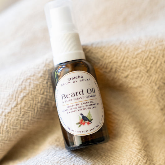 A bottle of Grateful Glow by Becky Beard Oil resting on a textured beige woven blanket. The amber glass bottle has a white spray cap and a white label with black text and a small botanical illustration.