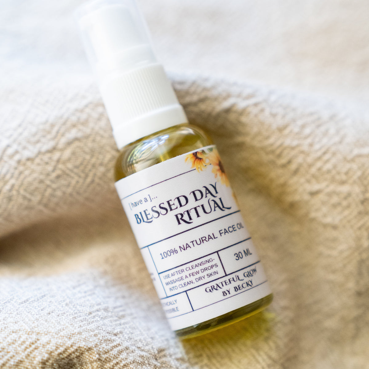 A small bottle of face oil with a white spray top and white label reading 'Blessed Day Ritual' and '100% Natural Face Oil,' placed on a beige textured fabric surface.