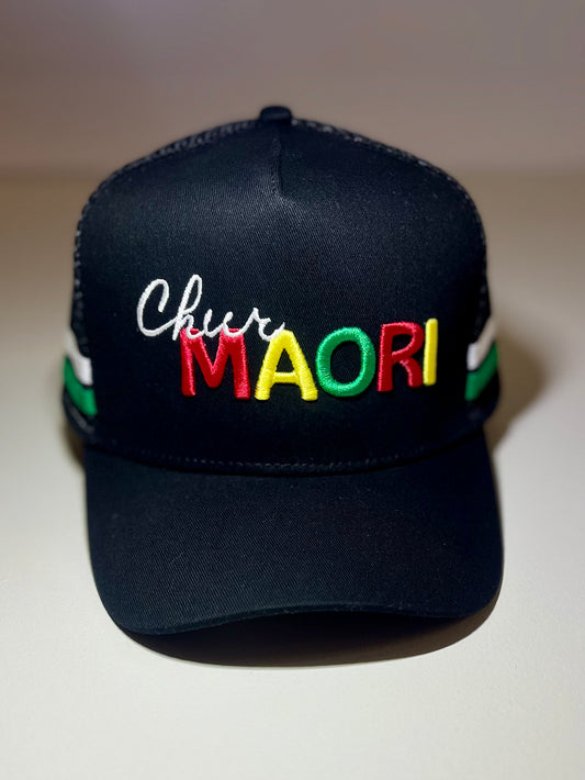 A black trucker-style baseball cap with 'Chur' in white script and 'MAORI' in large red, yellow, and green letters across the front. The cap has green and white striped mesh side panels and is photographed against a neutral beige background.
