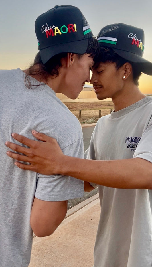 Two young men embracing outdoors at sunset, both wearing matching black baseball caps with 'Chur Maori' embroidered on the front in red, green, yellow, and black.