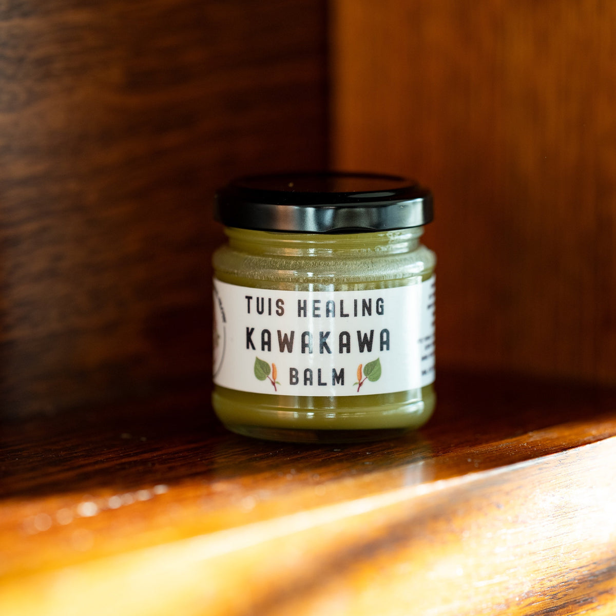 A small glass jar of Tuis Healing Kawekawa Balm with a black lid and white label, placed on a warm wooden surface with a blurred wooden background.