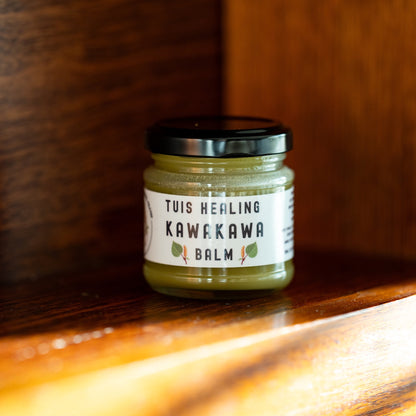 A small glass jar of Tuis Healing Kawekawa Balm with a black lid and white label, placed on a warm wooden surface with a blurred wooden background.