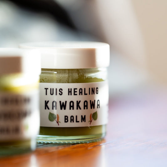 A close-up of a small glass jar labeled 'TUIS HEALING KAWAKAWA BALM' with a white lid, sitting on a wooden surface with another blurred jar beside it.