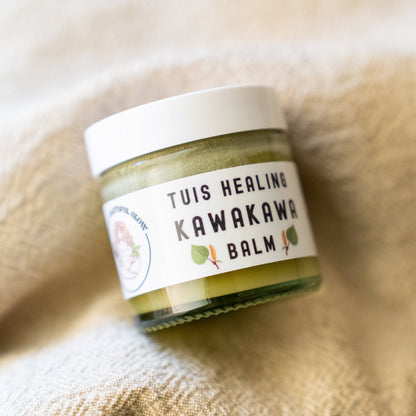 A small glass jar of Tuis Healing Kawakawa Balm with a white lid and white label, resting on a soft beige textured blanket.