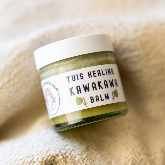 A small glass jar of Tuis Healing Kawakawa Balm with a white lid and white label, resting on a soft beige textured blanket.