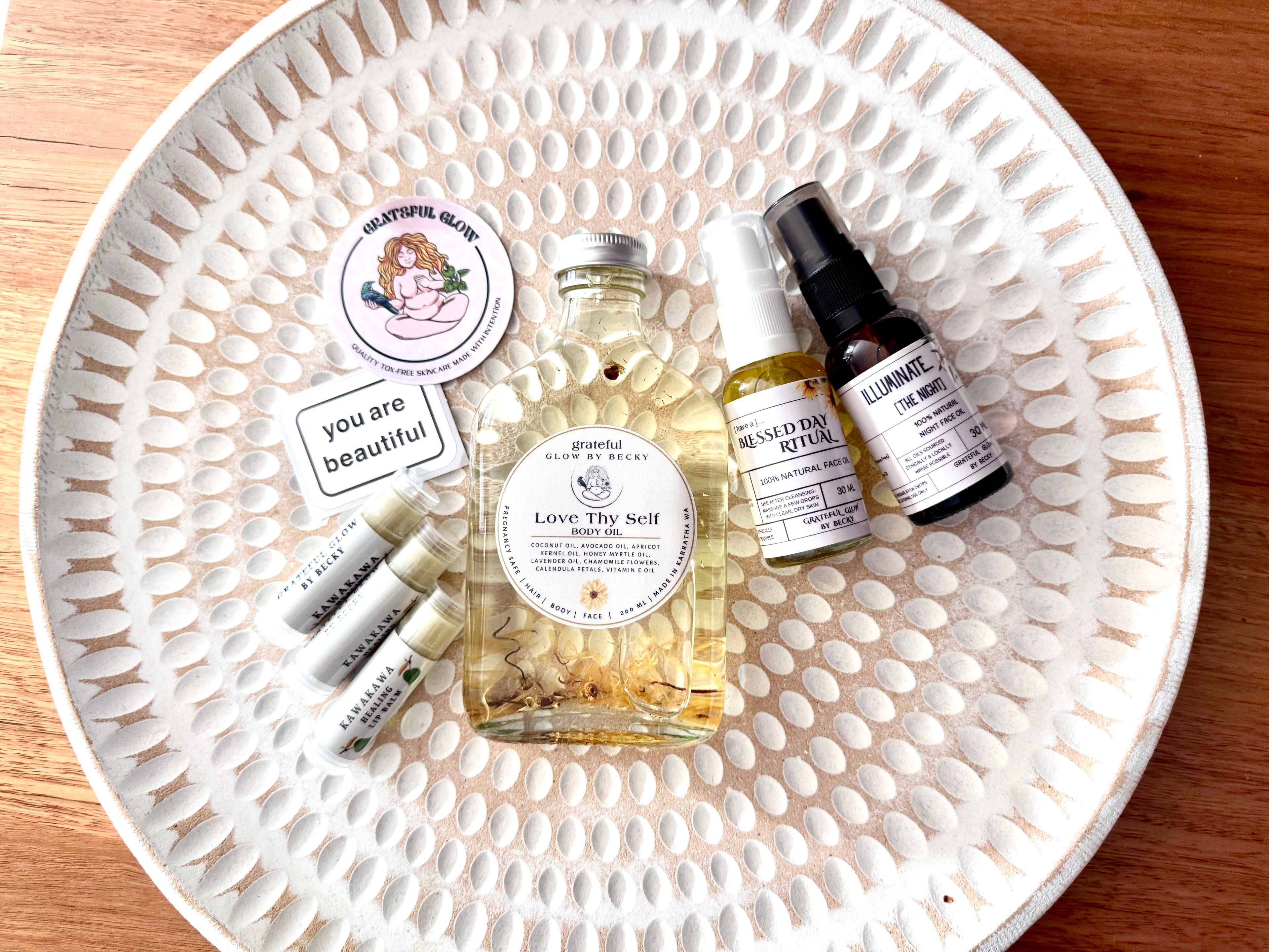 A curated collection of wellness products arranged on a white textured ceramic plate, including a central bottle of Love Thy Self body oil, two small bottles of face oil, gratitude art stickers, and three lip balm tubes, photographed from above on a wooden surface.