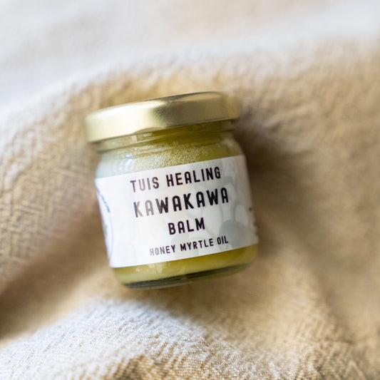 A small glass jar of Tuis Healing Kawakawa Balm with Honey Myrtle Oil with a gold lid and white label, resting on a textured beige fabric background.
