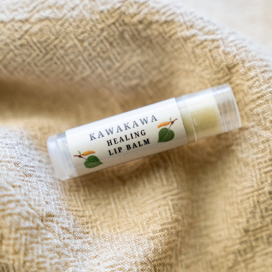 A clear lip balm tube labeled 'KAWAKAWA HEALING LIP BALM' resting on a textured beige woven fabric background.