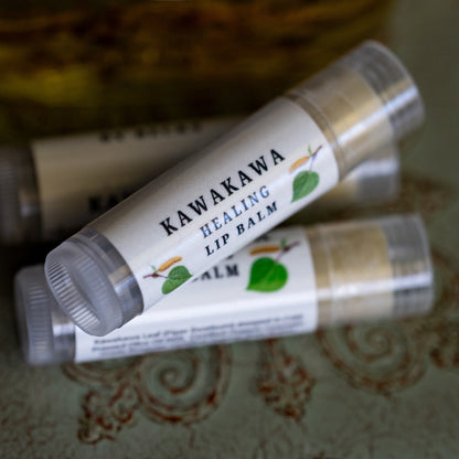 Two lip balm tubes labeled 'KAWAKAWA HEALING LIP Balm' resting on a patterned green surface, with a large bottle of 'Love Thy Self' body oil in the background.