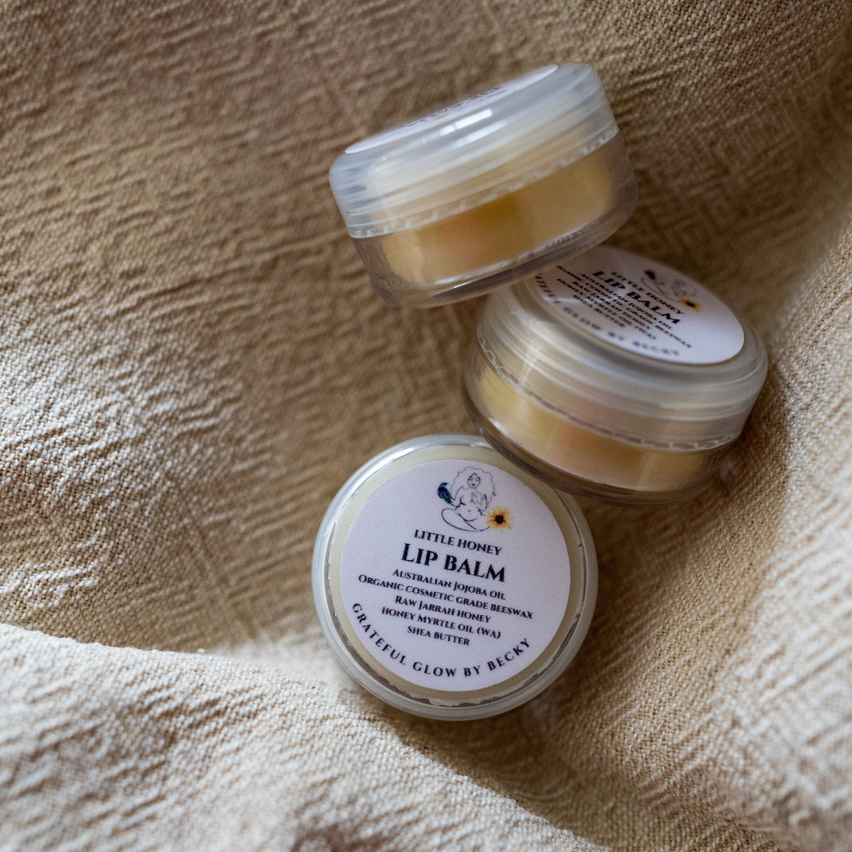 Three small round jars of lip balm arranged on a textured beige fabric background.