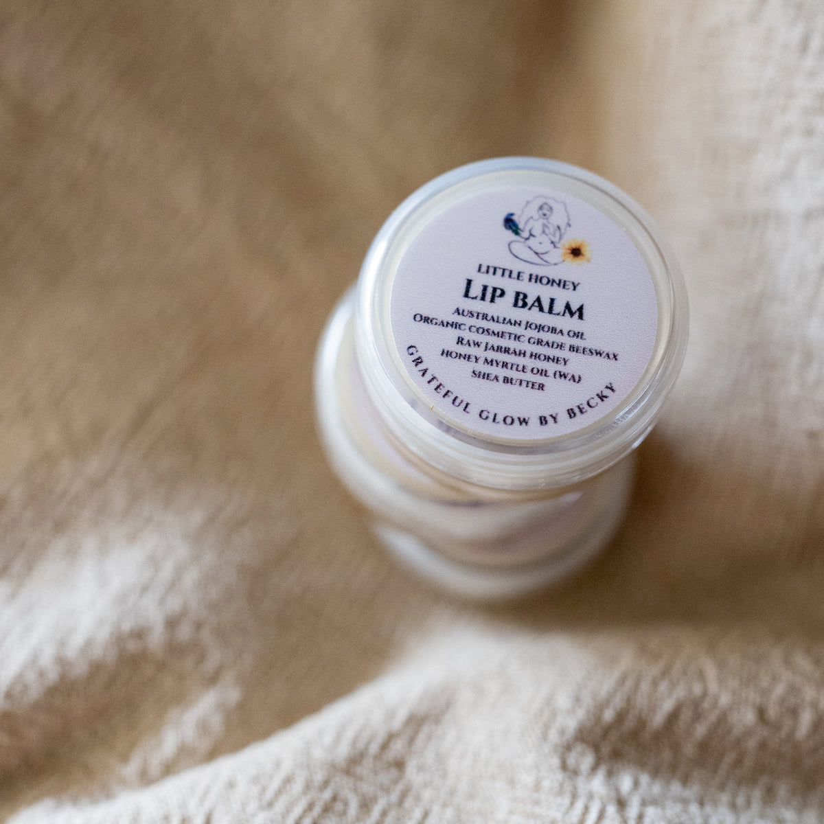 A small jar of lip balm with a white lid labeled 'Little Honey Lip Balm' resting on a soft beige textured fabric background.