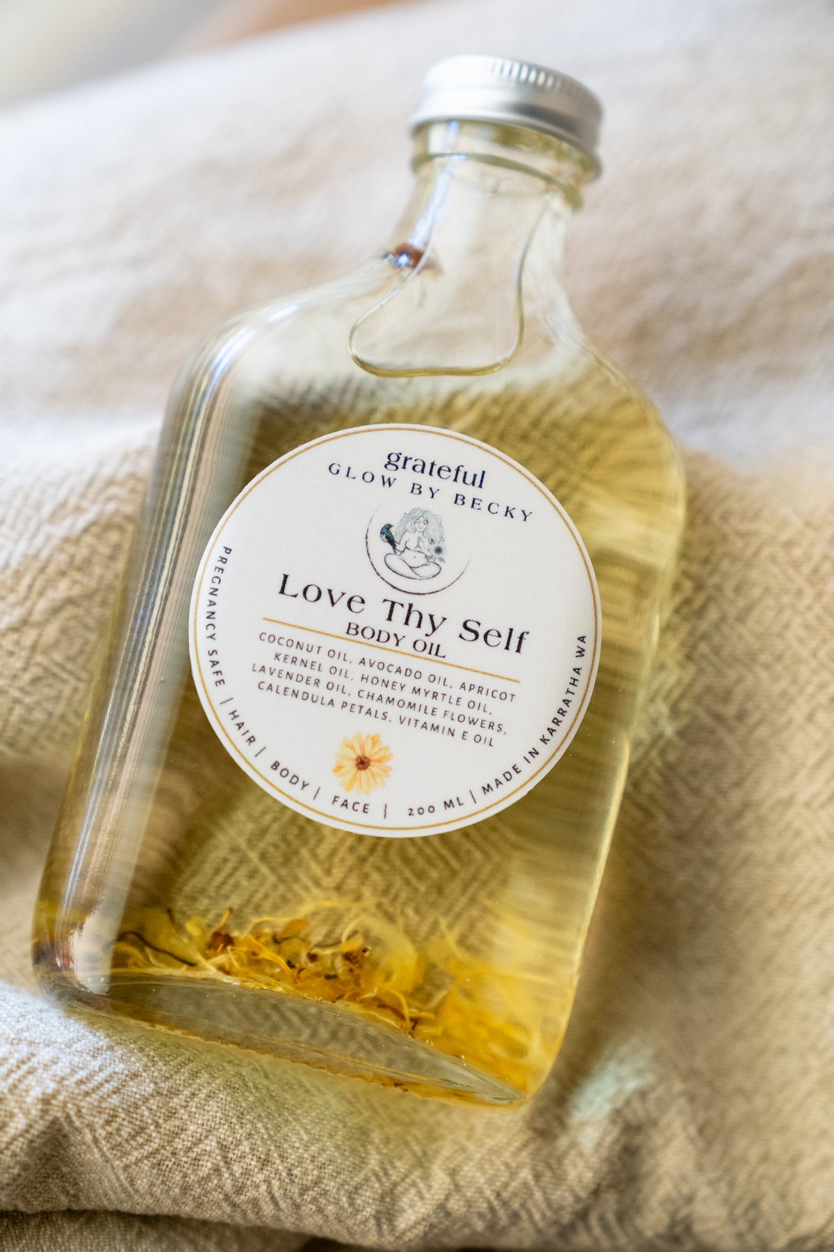 A clear glass bottle of Grateful Glow by Becky Love Thy Self Body Oil filled with pale yellow oil and dried yellow flowers, resting on a textured beige fabric background.
