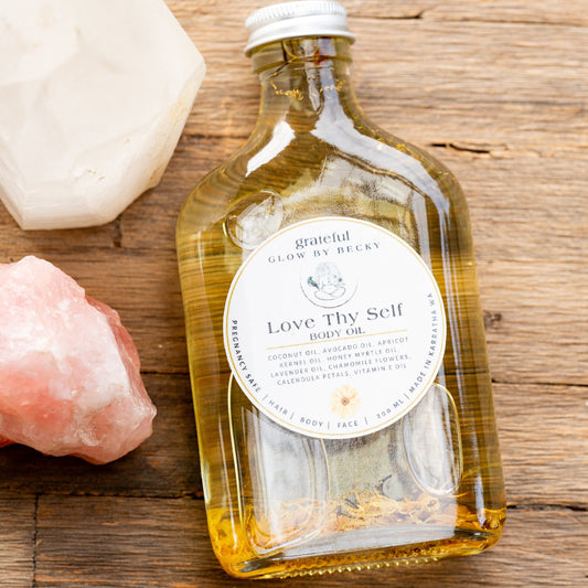 A glass bottle of 'Love Thy Self' body oil by Grateful Glow by Becky, filled with golden-yellow oil, displayed on a rustic wooden surface alongside a white crystal and a pink crystal.