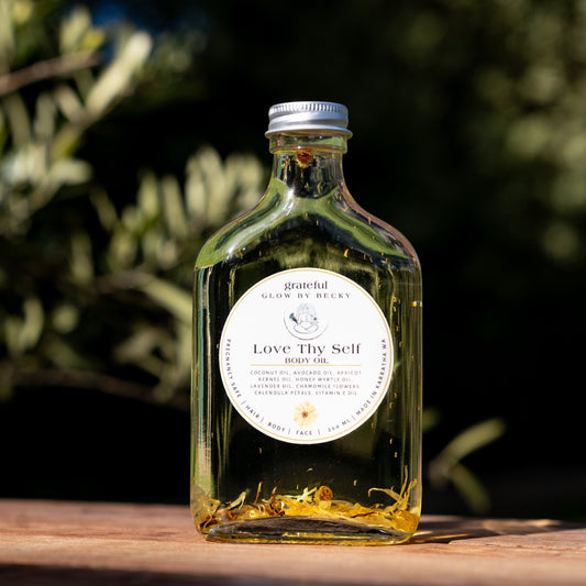 A clear glass bottle of Love Thy Self Honey Oil with a white label and silver cap, sitting on a wooden surface with a blurred outdoor garden background.