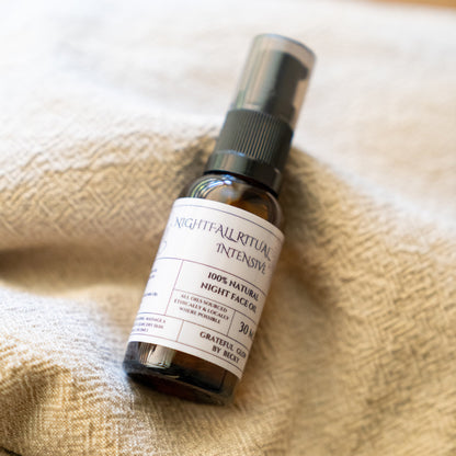 A small amber glass spray bottle labeled 'Nightfall Ritual Intensive' and '100% Natural Night Face Oil' resting on a textured beige woven blanket.