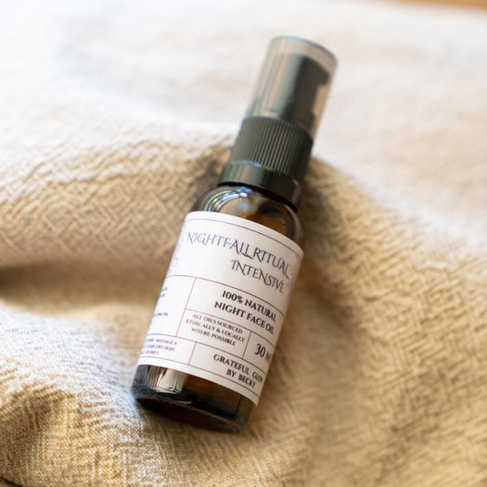 A small amber glass spray bottle labeled 'Nightfall Ritual Intensive' and '100% Natural Night Face Oil' resting on a textured beige woven blanket.